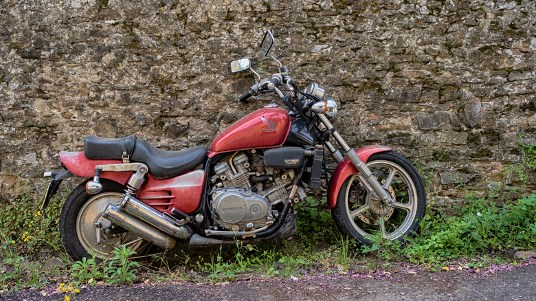 A red and black Honda Magna VF750C parked next to a stone wall on grass at the side of a road.