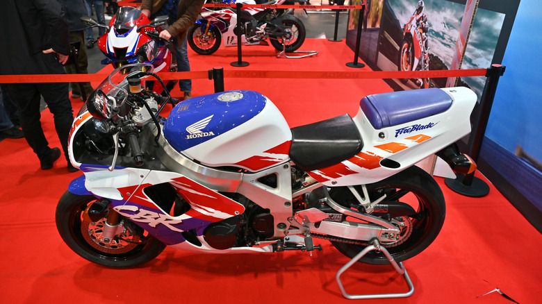 A white, red, and blue 1992 debut Honda CBR900RR Fireblade motorbike parked indoors at an exhibition on red carpet.