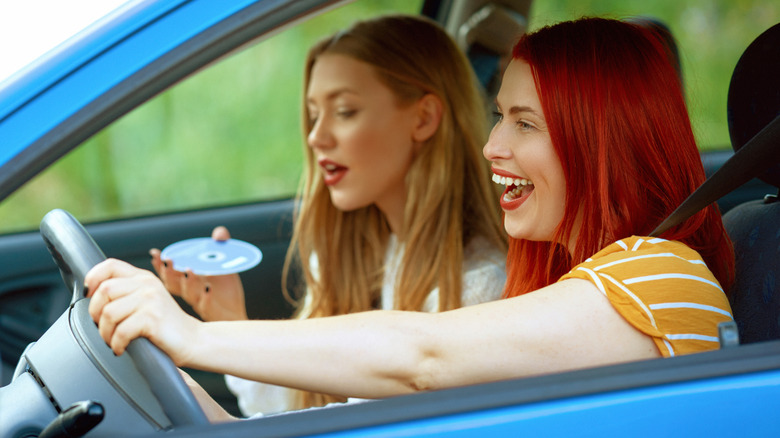 A driver smiling and singing along to music while the passenger sings and changes CDs in the car