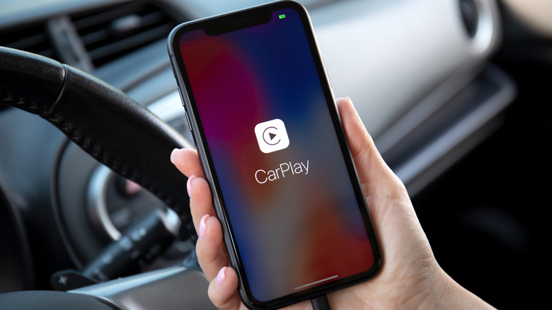 Hand holding iPhone in front of auto steering wheel with CarPlay logo on screen.