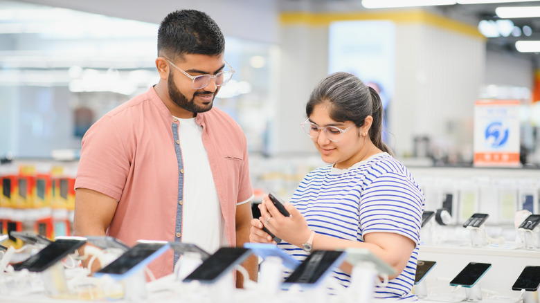 A dark-haired man in a pink shirt and a dark-haired woman in a blue and white striped shirt look at a phone in a store
