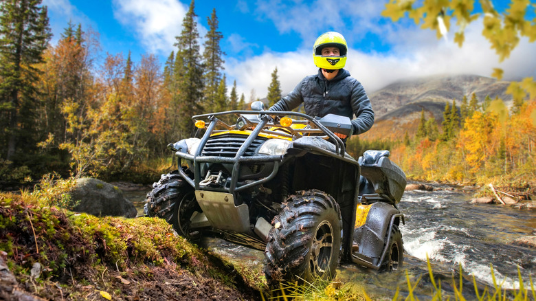 A man in a bright yellow helmet riding an ATV in the woods near a river