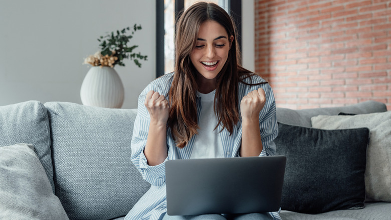 A woman who looks excited sitting on a sofa while holding a laptop on her laps
