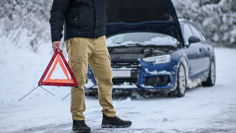 Man holding a warning triangle next to his broken down car on a snowy winter road.