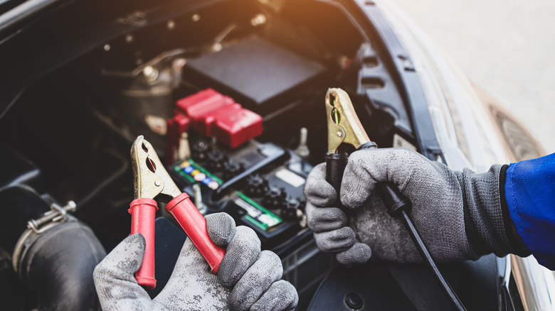 A pair of gloved hands holding red and black cable connectors near an open car hood
