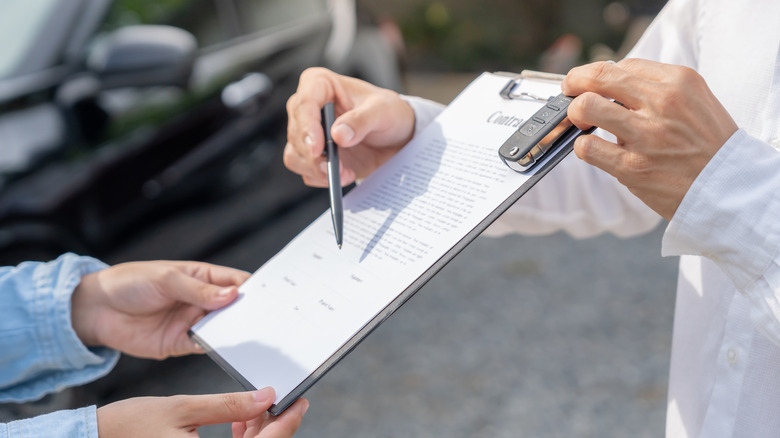 A close up of one hand holding a clipboard with paperwork while another set of hands holds out a pen and a car key fob