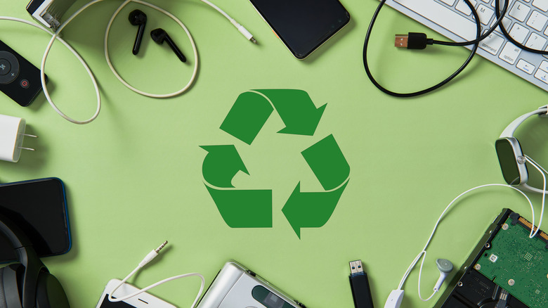 Phone, smartwatch, earbuds, cables, and computer components on green table with recycling symbol