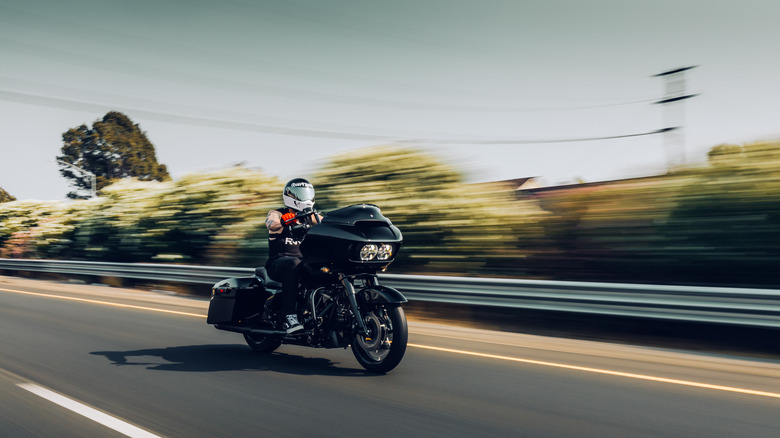 Person riding a Harley cruiser along a highway. The bike is in focus, while the background is blurred.