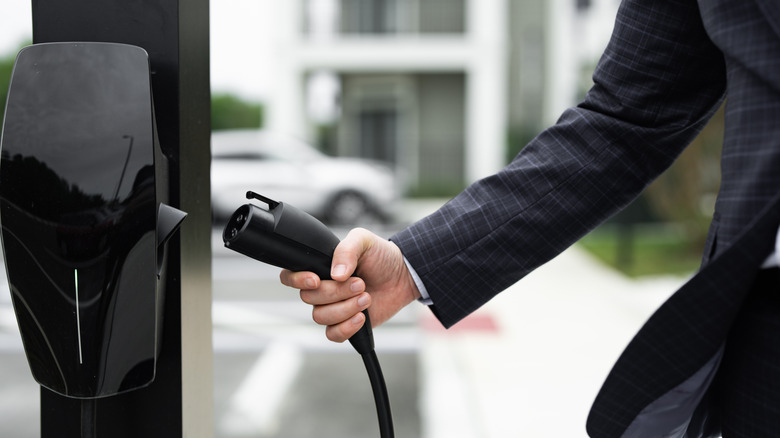 A man in a dark suit holding a plug at a public EV charging station in a parking lot