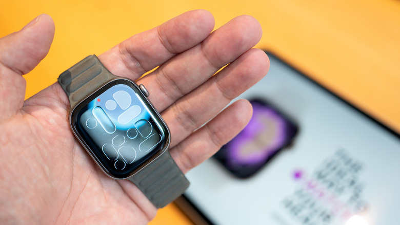 Person holding an Apple Watch in their palm with a blurry iPad in the background